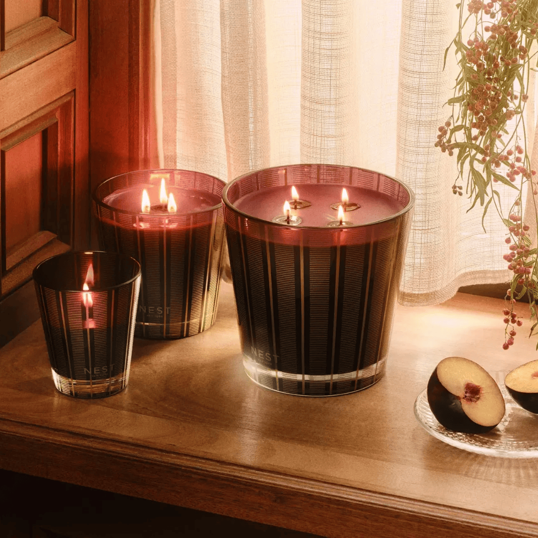 Three lit NEST candles in ribbed glass holders arranged on a wooden table near a window with sheer curtains, with sliced fruit on a plate nearby.